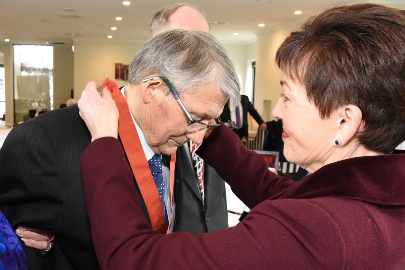 Image of Dame Patsy placing the insignia of a CNZM around Jim Anderton's neck